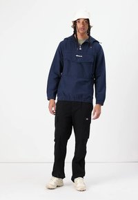 Blå genserjakke med lomme foran, elastisk kant og hette med snor, kombinert med svarte cargobukser og lyse sneakers.