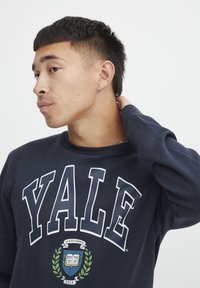 Felpa blu navy con la scritta "YALE" in bianco, accompagnata da un emblema universitario e un design a alloro al centro. Tessuto morbido e maniche lunghe.
