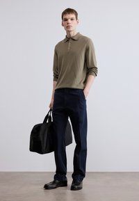 Πράσινο ελιάς πόλο με μακριά μανίκια και κουμπιά, σε συνδυασμό με σκούρο ναυτικό παντελόνι και μαύρα παπούτσια. Το μοντέλο κρατά μια μαύρη τσάντα.