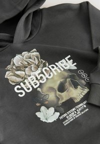Hoodie noir avec un graphique floral et crânien, portant le texte blanc "SUB5CRIBE". En matériau coton, texture douce et poignets côtelés. Éléments de design artistique.
