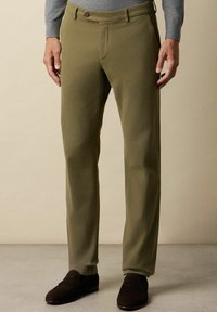 Oliwkowe spodnie bawełniane o prostym kroju, z pojedynczym zapięciem na guzik z przodu i kieszeniami. Noszone z ciemnobrązowymi butami.