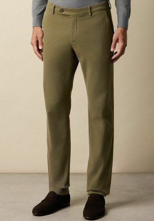 Olijfgroene katoen broek met een rechte snit, voorzien van een enkele knoopsluiting aan de voorkant en zakken. Draagt bij donkere bruine schoenen.