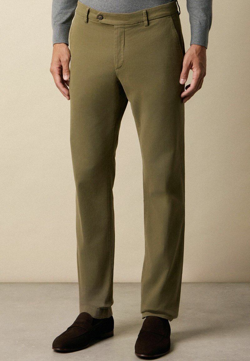 Oliwkowe spodnie bawełniane o prostym kroju, z pojedynczym zapięciem na guzik z przodu i kieszeniami. Noszone z ciemnobrązowymi butami.
