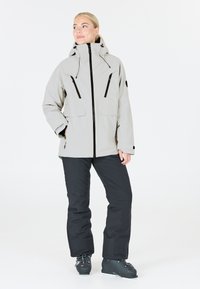 Lichtgrijze waterdichte jas met hoge kraag, zwarte rits en zakken; gecombineerd met zwarte sneeuwbroeken en zwarte skibotten.
