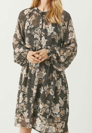Femme portant une robe noire à manches longues avec des motifs floraux beige et blanc, debout, les mains jointes devant elle.