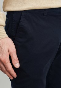 Chino blu navy realizzati in tessuto di cotone, caratterizzati da una consistenza liscia e tasche laterali. Mano appoggiata sulla coscia, con un maglione di lana beige neutro.