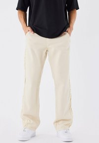 Pantaloni beige a gamba larga con cuciture laterali a taglio vivo, realizzati in tessuto denim. Abbinati a sneakers bianche e una maglia nera.
