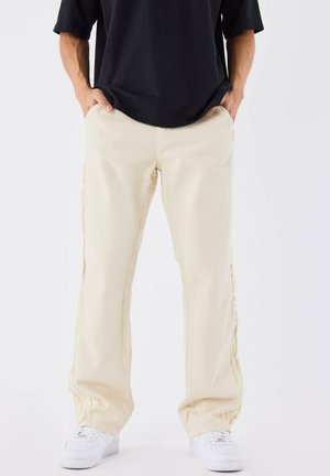 Pantaloni beige a gamba larga con cuciture laterali a taglio vivo, realizzati in tessuto denim. Abbinati a sneakers bianche e una maglia nera.