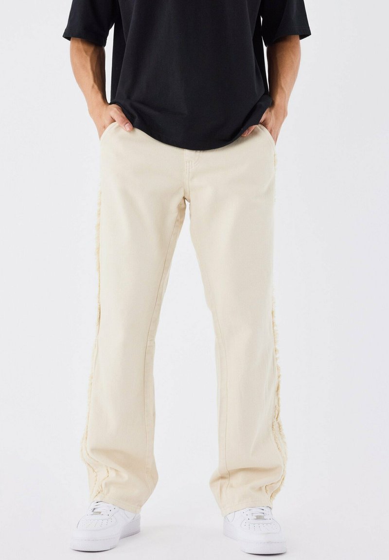 Pantaloni beige a gamba larga con cuciture laterali a taglio vivo, realizzati in tessuto denim. Abbinati a sneakers bianche e una maglia nera.