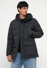 Puffer jacket in dark gray with a hood, front zipper, and two large pockets. Features quilted design and adjustable cuffs for warmth.