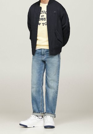 Persona con chaqueta bomber negra, sudadera gráfica color crema, vaqueros azul claro y zapatillas blancas de pie con las manos detrás de la espalda.