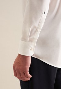 Männers Hand neben weißem Langarmhemd mit Knöpfen und kleinem besticktem Detail einer schwarzen und roten Rose, trägt schwarze Hose.