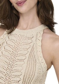 Haut halter en tricot beige avec un design en dentelle ajourée, un col montant et des détails à volants sur les épaules. Le fil texturé ajoute de la dimension.