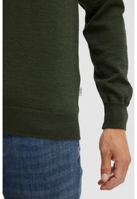 Suéter de punto verde oscuro con un acabado texturizado, con un dobladillo y puños acanalados. Una pequeña etiqueta es visible cerca del dobladillo. Combinado con unos vaqueros azules.