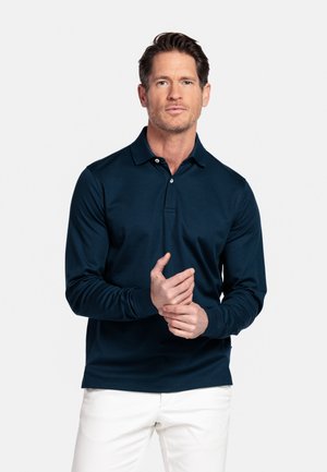 Man met kort bruin haar, gekleed in een marineblauw poloshirt met lange mouwen en een witte broek, staand tegen een effen witte achtergrond, waarbij hij zijn linkerhand aanraakt.