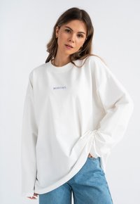 Witte longsleeve sweatshirt met "MIRONS" geborduurd in lichtblauw. Zachte stof, ontspannen pasvorm, afgeronde zoom, gecombineerd met blauwe jeans.