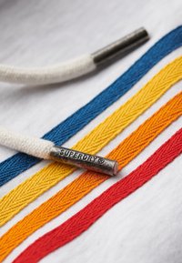 Biała tkanina z pięcioma teksturowanymi paskami w kolorach niebieskim, żółtym, pomarańczowym, czerwonym oraz białą koronką. Srebrna metalowa końcówka sznurka jest ozdobiona napisem „SUPERDRY”.