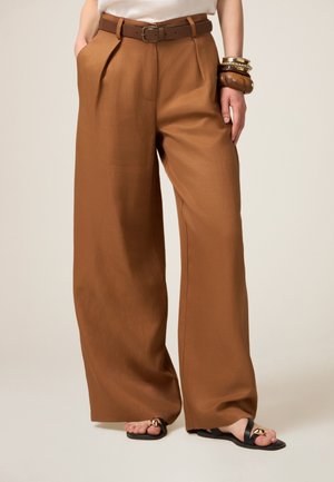Wijde broek in karamelkleurige stof, met voorplooien en een bruine riem. Gecombineerd met eenvoudige zwarte sandalen en gestapelde armbanden.