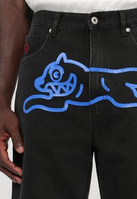 Jeans de mezclilla negros con un gran gráfico azul de un gato estilizado en la pierna izquierda, y detalles en rojo en el bolsillo.