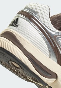 Sportovní obuv s texturovaným stříbrným a béžovým svrškem, hnědými akcenty a černou podešví s kulatým výřezem a vzorovaným profilem.