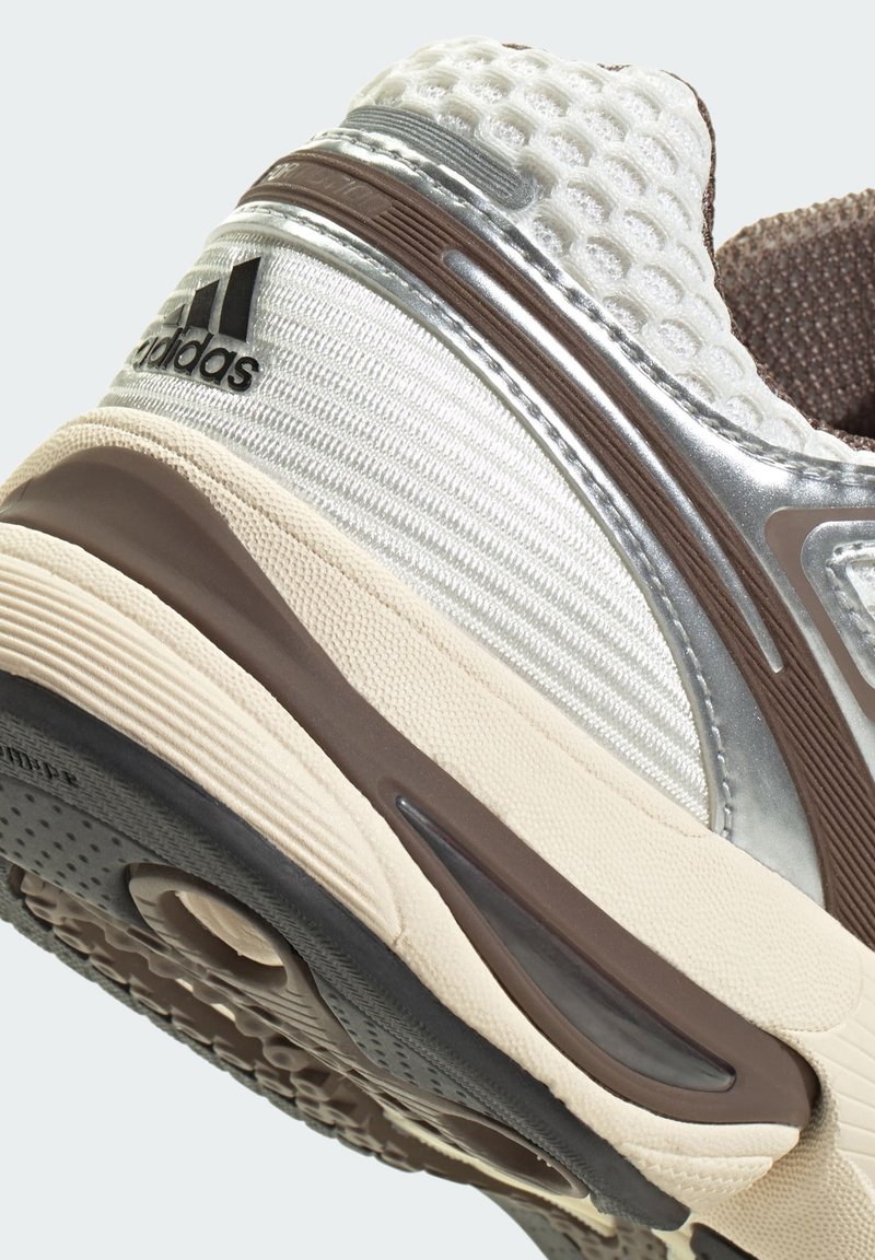 Sportschuh mit einer strukturierten silbernen und beigen Oberseite, braunen Akzenten und einer schwarzen Laufsohle mit einem runden Ausschnitt und strukturiertem Profil.