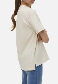 T-shirt à manches courtes et coupe décontractée en coton beige clair, avec des fentes latérales et un col rond, associé à un jean bleu.