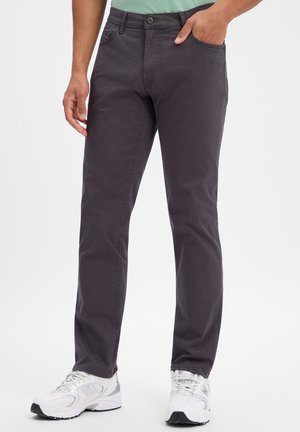 Personne portant un pantalon gris foncé droit et des baskets blanches, debout, une main dans la poche, sur un fond uni.