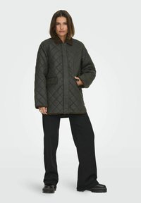 Giacca trapuntata verde con colletto marrone, tasche frontali e chiusura con zip. Abbinata a pantaloni neri e stivaletti con tacco.