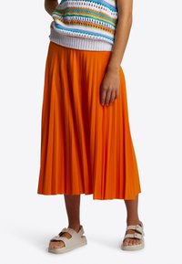 Oranssi, midimittainen pliseerattu hame, jossa sileä tekstuuri, yhdistettynä valkoisiin paksupohjaisiin sandaaleihin, joissa on kaksi säädettävää solkea.