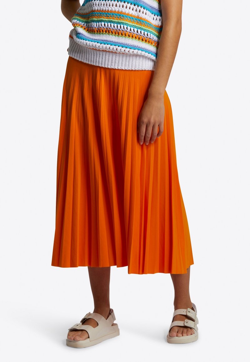 Oranssi, midimittainen pliseerattu hame, jossa sileä tekstuuri, yhdistettynä valkoisiin paksupohjaisiin sandaaleihin, joissa on kaksi säädettävää solkea.