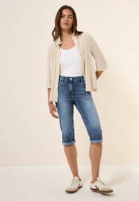 Camisa de botones beige claro sobre una camiseta blanca, combinada con capris de mezclilla azul y zapatillas blancas con acentos rojos. Textura de tela suave.