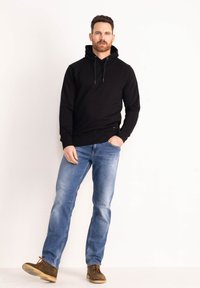 Felpa nera in cotone con cappuccio e lacci, abbinata a jeans in denim azzurro chiaro. Il modello indossa scarpe in camoscio marroni. Un outfit semplice e casual.