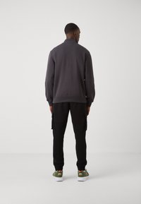 Sudadera gris con un cuello alto, combinada con pantalones cargo negros y zapatillas deportivas verdes con acentos naranjas. Textura de tela suave.