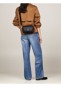 Musta nahkainen olkalaukku, jossa on hopeanväriset yksityiskohdat, suorakaiteen muotoinen ja sileä pinta, käytetty ruskean takin ja sinisten farkkujen kanssa.