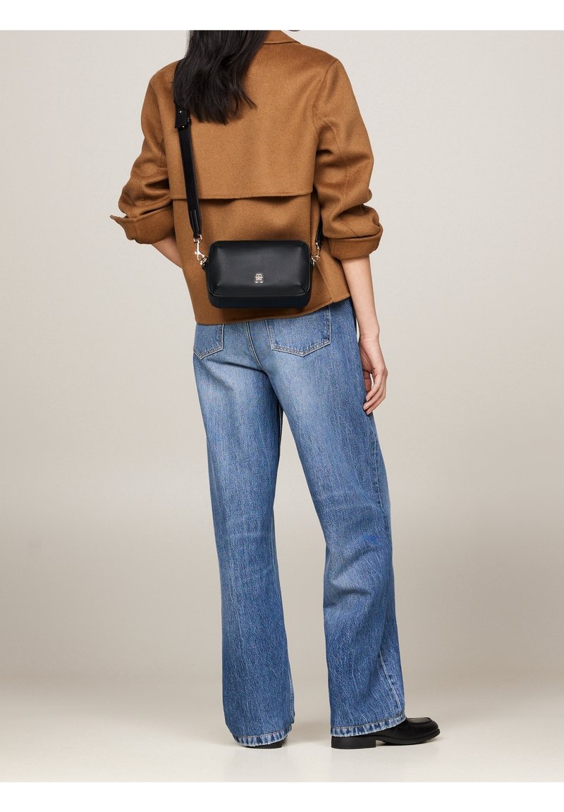 Musta nahkainen olkalaukku, jossa on hopeanväriset yksityiskohdat, suorakaiteen muotoinen ja sileä pinta, käytetty ruskean takin ja sinisten farkkujen kanssa.