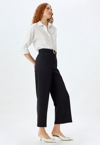 Chemise blanche à manches retroussées, pantalon noir taille haute avec une ceinture, et chaussures à talons blanches. Design épuré, silhouette ajustée.