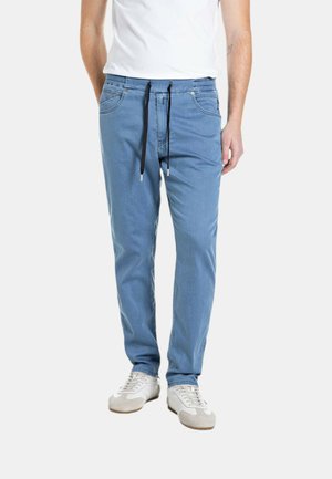 Uomo che indossa pantaloni jogger di denim azzurro chiaro con coulisse nere, sneakers bianche e t-shirt bianca, in piedi davanti a uno sfondo uniforme.