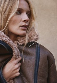 Bruine leren jas met een zachte crèmekleurige kraag, voorzien van een rits en een gestructureerd materiaal. Model grijpt de jas vast om het gedetailleerde ontwerp te tonen.