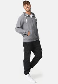 Sweat à capuche gris zippé avec cordons, poches latérales, associé à un pantalon cargo noir avec poches, baskets blanches. Esthétique décontractée et confortable.
