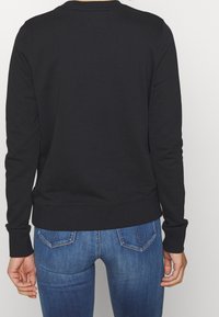 Persona con un sudadera negra de manga larga y jeans de mezclilla azules, vista desde atrás sobre un fondo claro y liso.