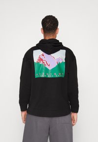 Sudadera negra con un gráfico de montaña colorido en la parte posterior, con acentos de texto en rosa, verde y rojo que dice "BASECAMP SIGUE EL CAMINO."