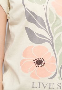 T-shirt beige clair avec impression florale. Caractéristiques d'une grande fleur pêche avec un centre noir, des feuilles vertes, et le texte "LIVE SY." Texture de tissu lisse.