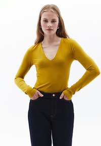 Top a maniche lunghe giallo senape con scollatura a V profonda. Realizzato in un tessuto morbido, presenta maniche aderenti e una texture liscia. Abbinato a jeans in denim scuro.