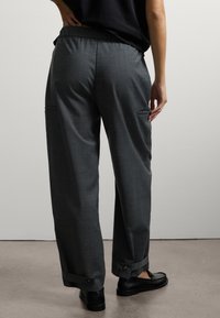 Persona con pantaloni larghi grigi scuri con fasce bottonate alle caviglie e mocassini neri, in piedi contro una parete leggera e semplice.