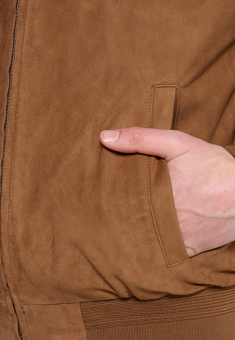 Ręka częściowo wsunięta w przednią kieszeń brązowej, zamszowej kurtki z widocznym zamkiem błyskawicznym i ściągaczem.
