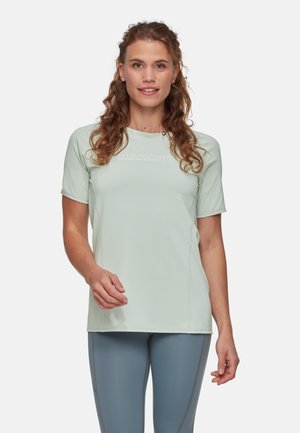 Vrouw met krullend haar, die een lichtgroen Mammut T-shirt en grijze legging draagt, staand tegen een effen witte achtergrond.