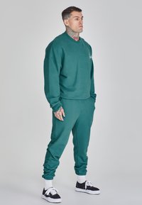 Sudadera en color verde azulado y pantalones de chándal a juego, fabricados en un tejido suave, con puños acanalados y un logotipo bordado en el pecho.