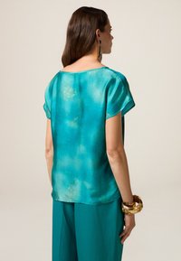 Top in seta a maniche corte di colore teal con un delicato motivo floreale. Vestibilità ampia e scollatura rotonda, abbinato a pantaloni coordinati e accessori dorati.