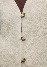 Gros plan d’un vêtement en polaire texturée couleur crème avec trois grands boutons ronds et un tissu marron en dessous.