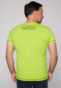 Camiseta de manga corta en un brillante color verde lima, con texto negro y símbolos náuticos en la parte posterior, y costuras contrastantes en negro.